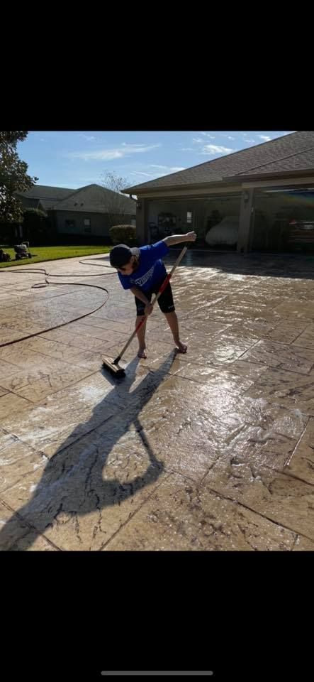A person washing a concrete driveway with a brush and water hose. Bright sunlight.