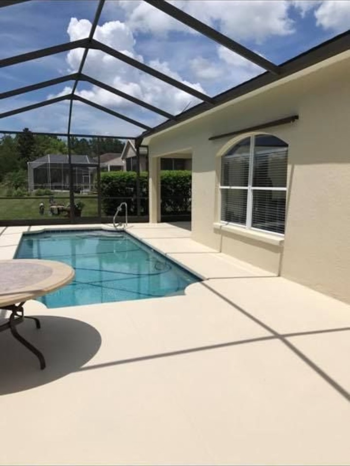 Pool deck with pool and screened enclosure, beige stucco house, blue sky with clouds.