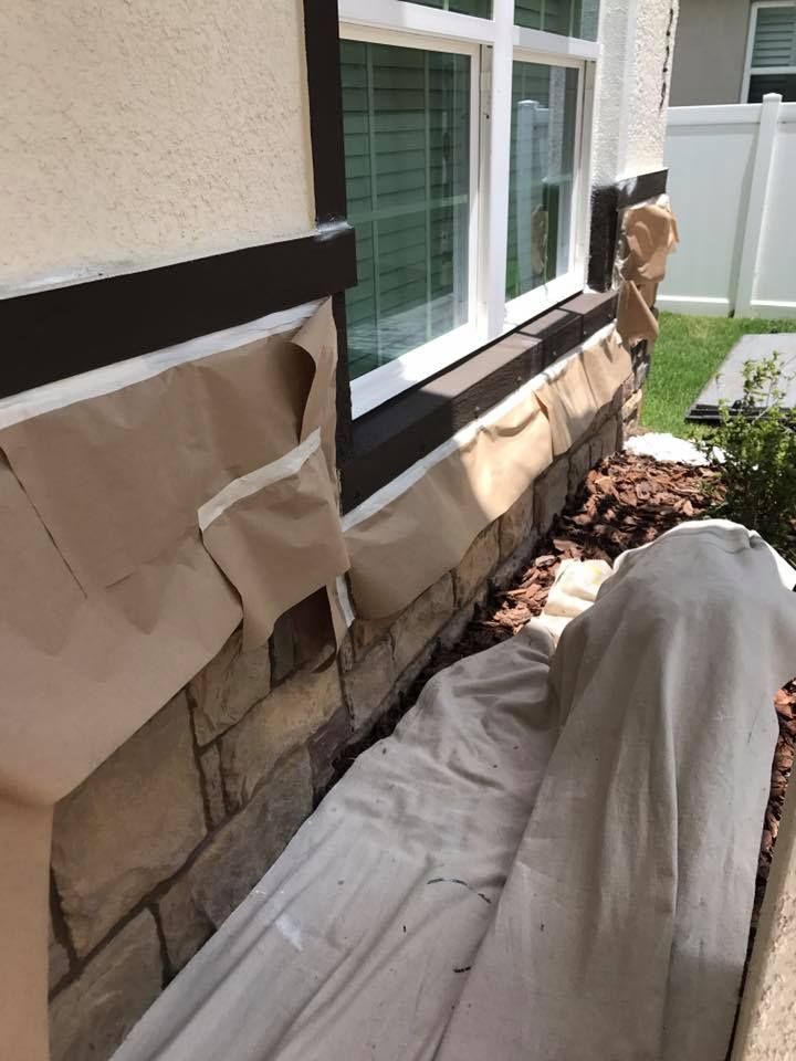 Exterior house being painted, masking tape and paper covering stone and trim, beige and brown colors.