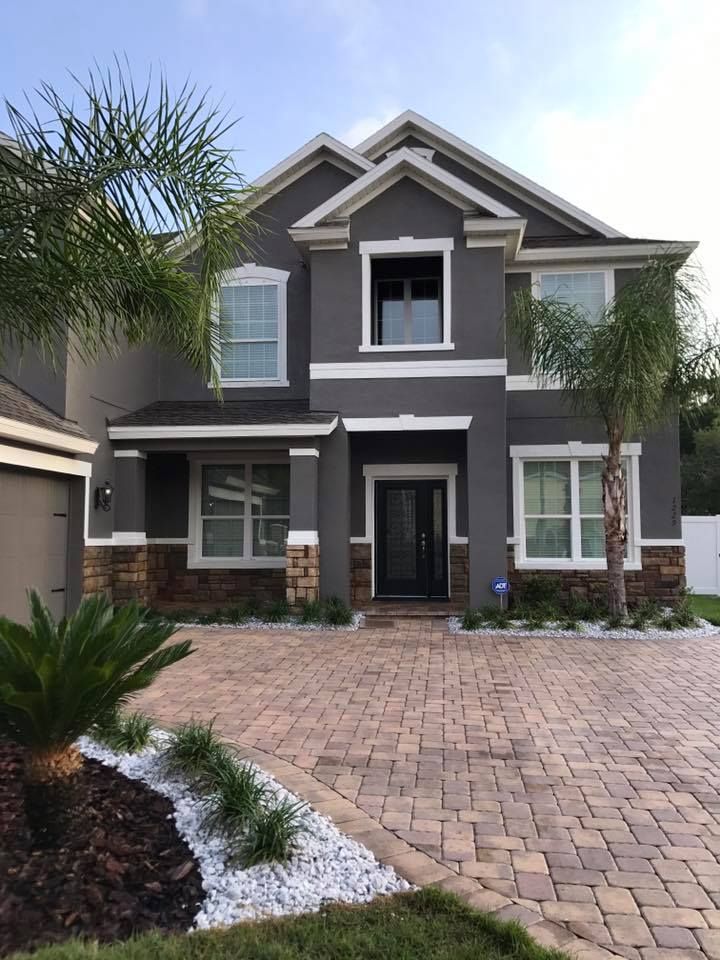 Two-story house with dark gray stucco, brown brick accents, white trim, and a brick driveway.