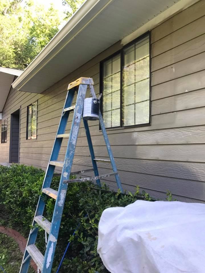A blue ladder with a paint can on the side of a house, covered in tan siding.