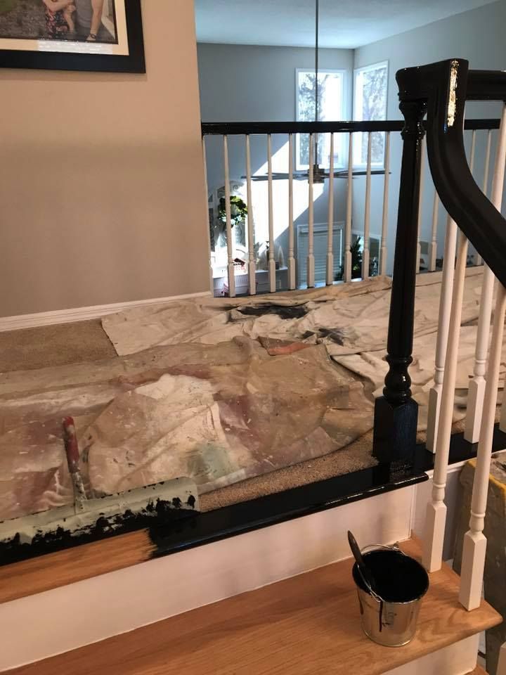 Staircase under renovation. Old carpet removed, exposed wood, black painted railing, white spindles, paint bucket on a step.