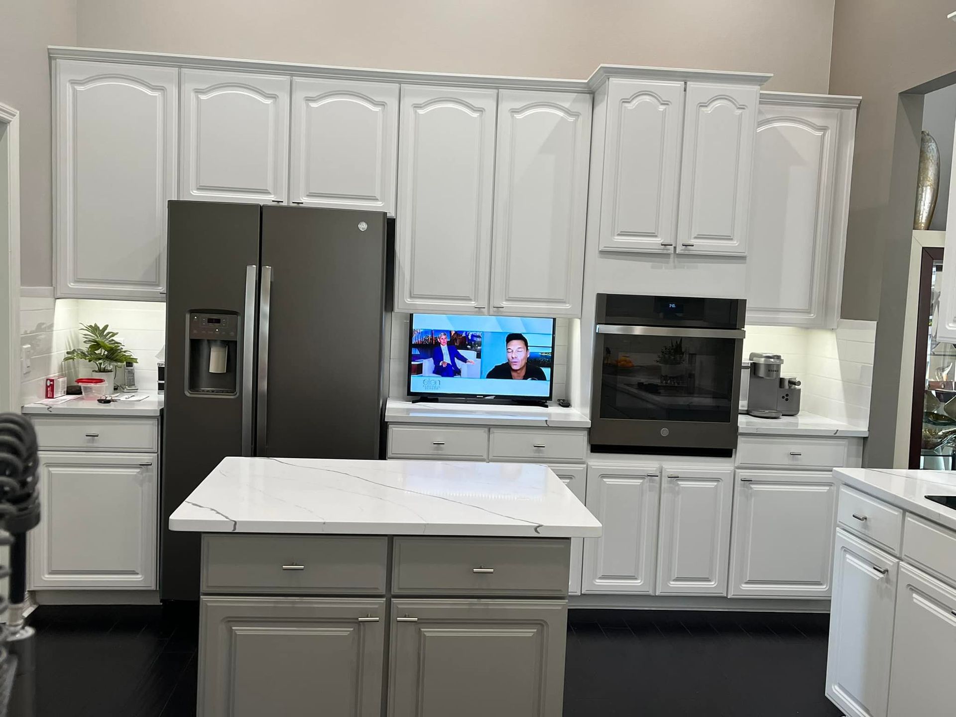 White kitchen cabinets with gray refrigerator, oven, and island. Television in center.