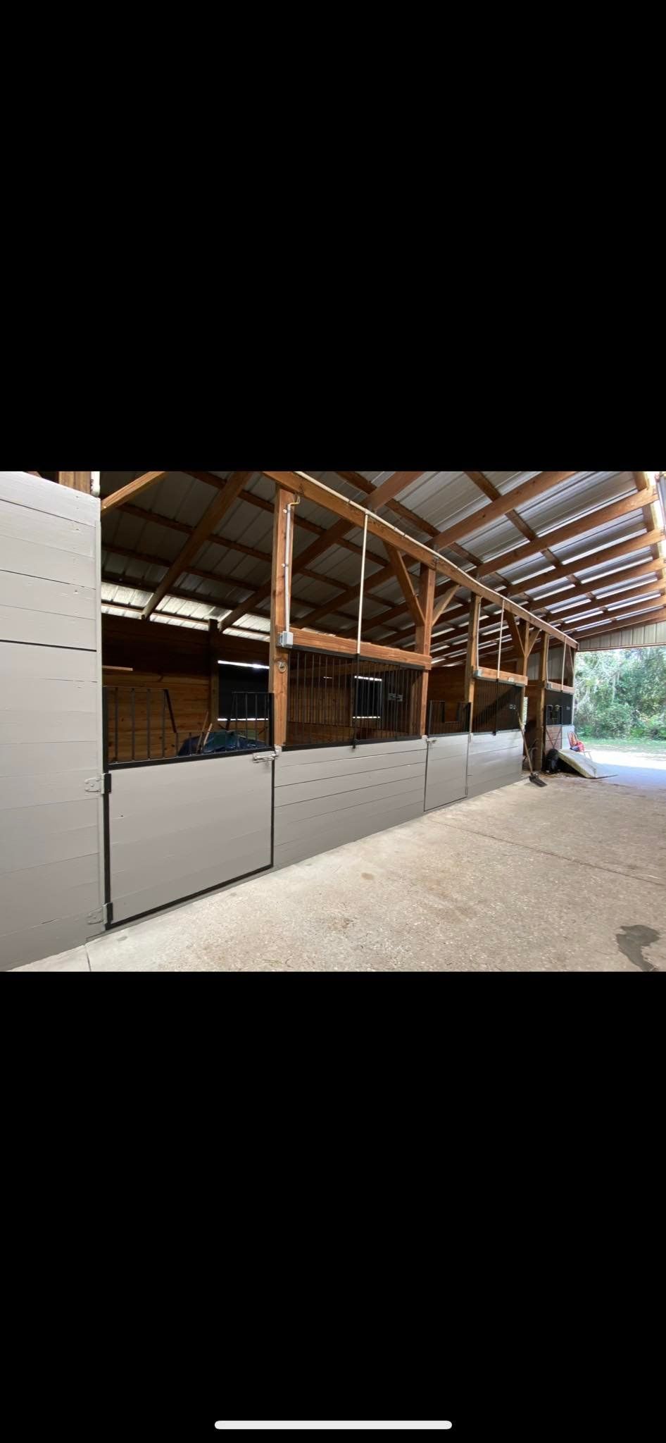 Horse barn interior, with stalls along the side. Wooden beams and corrugated metal roof. Light-colored walls.