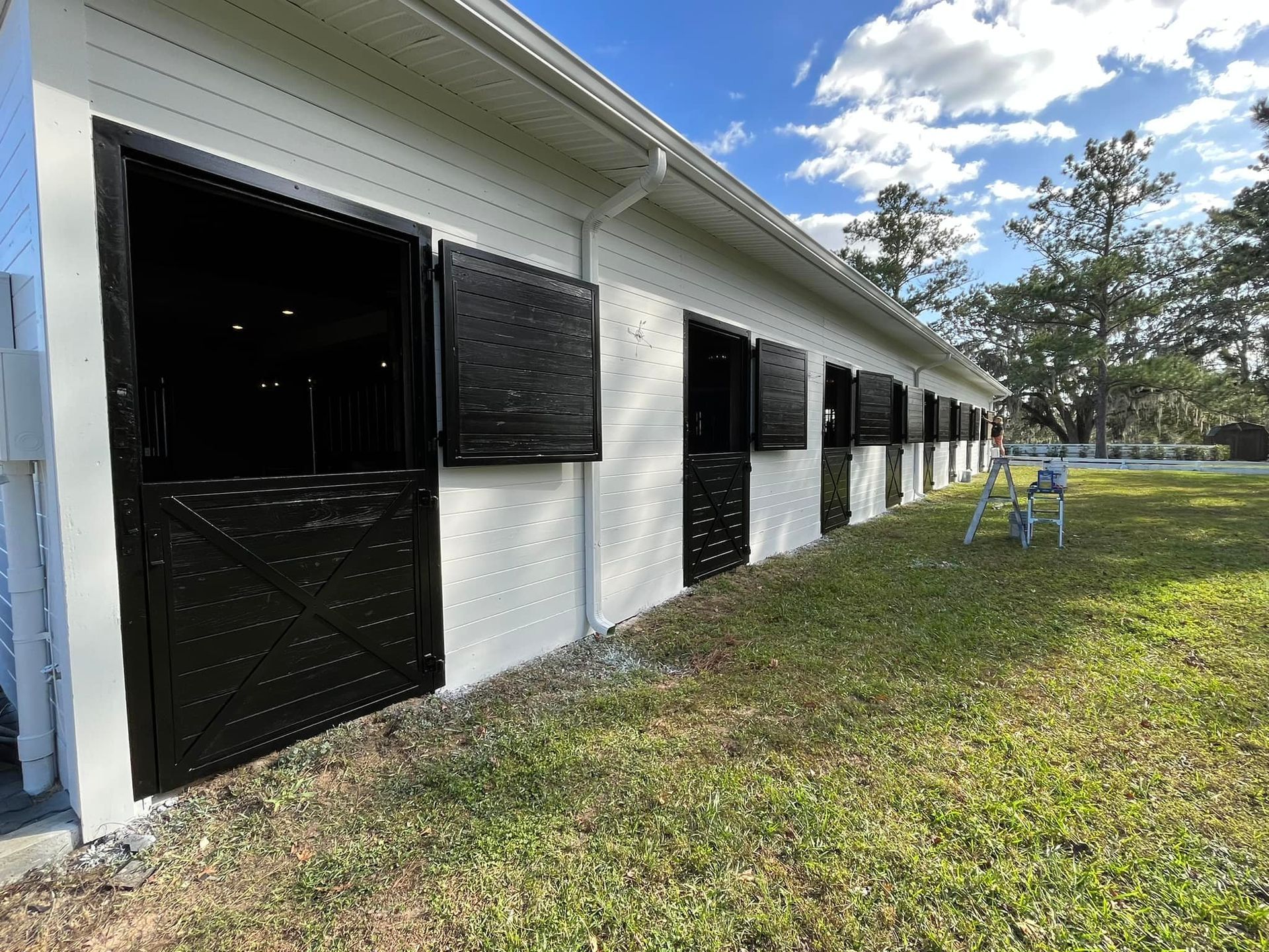 White horse stable with black doors and shutters, on a grassy field, under a blue sky.