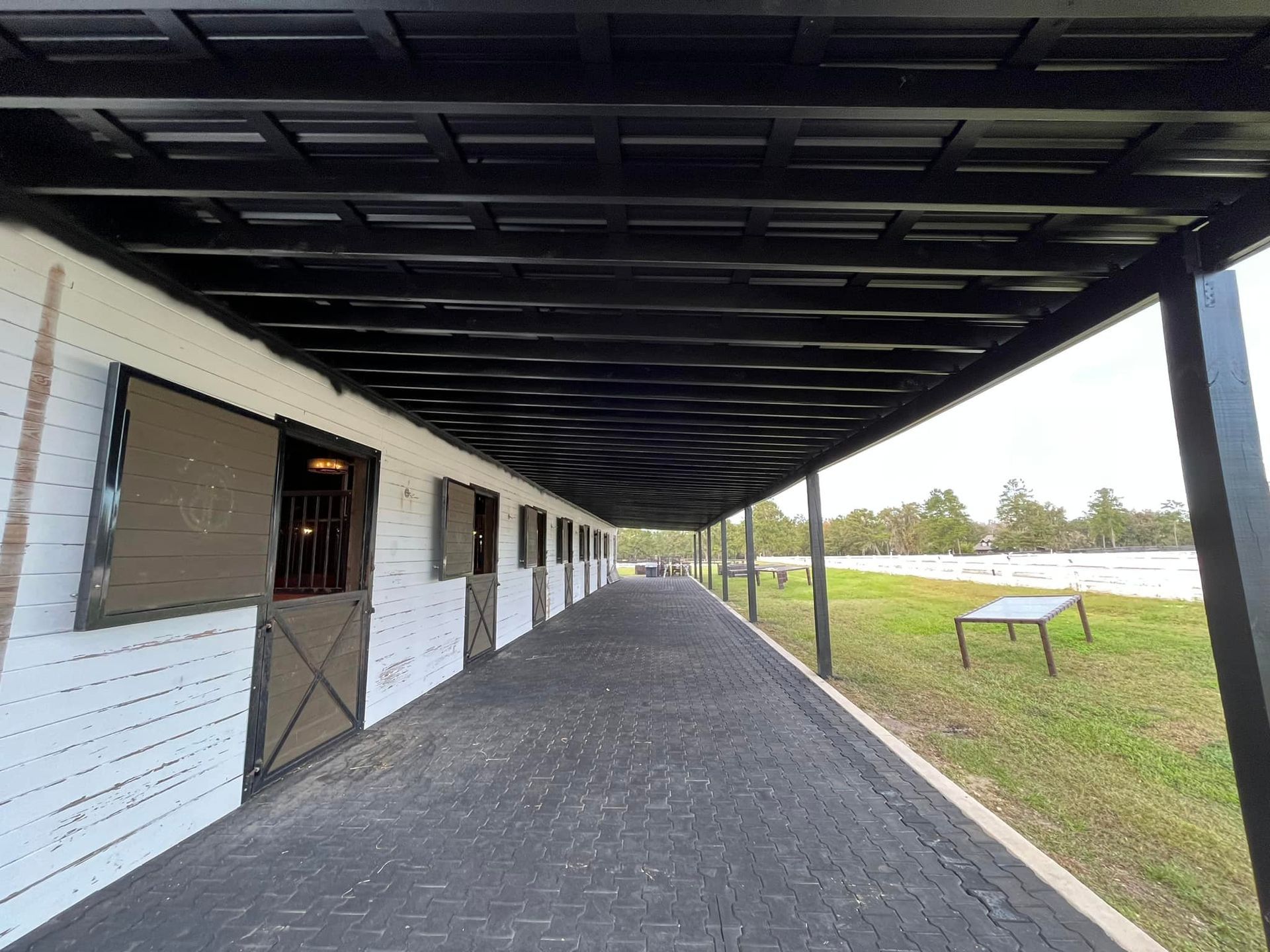 Black-roofed stable with white brick walls, dark doors, and a brick-paved walkway. A grassy field is on the right.