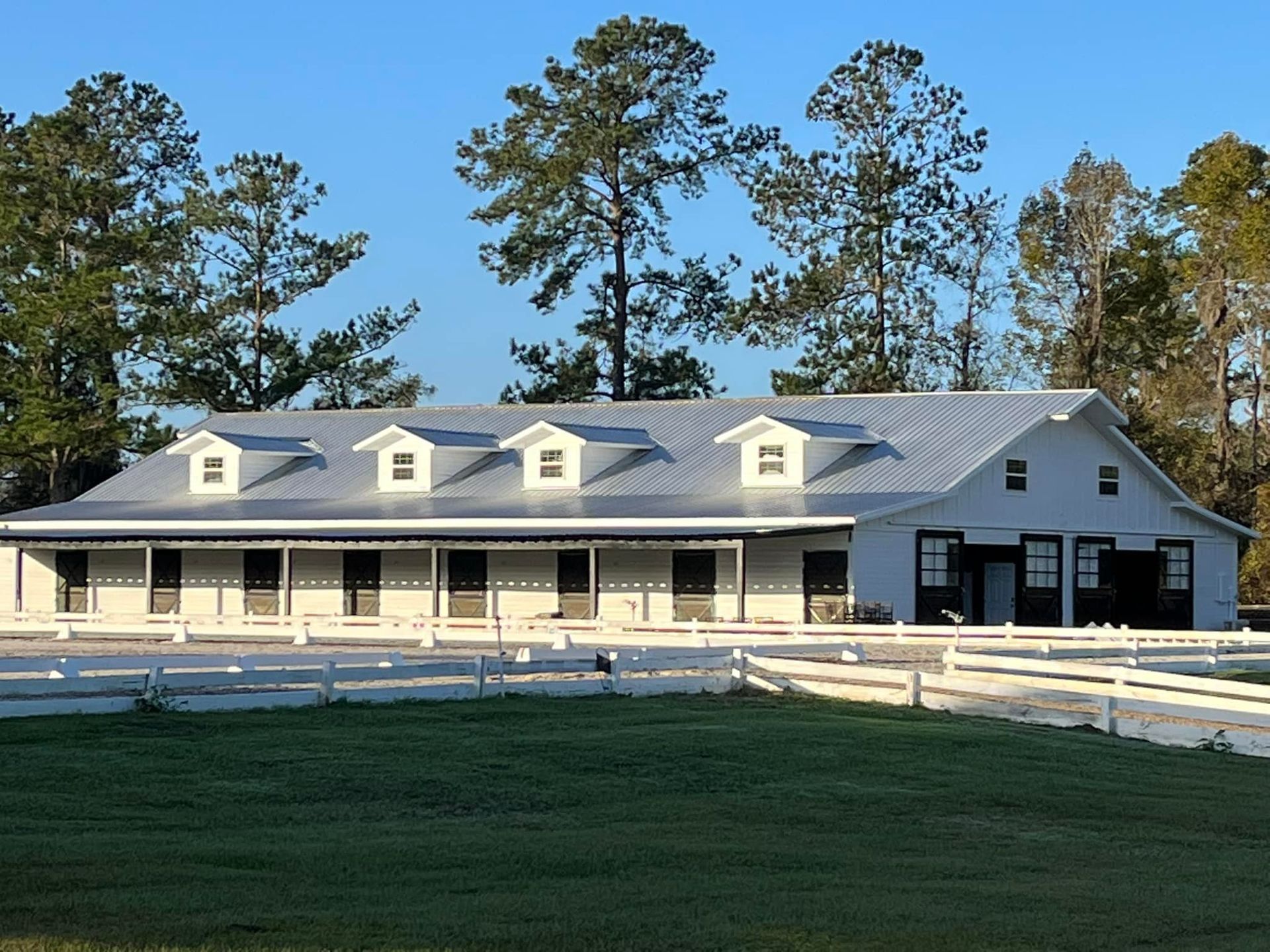 White horse stable with a silver roof and multiple windows, set in a grassy field with a white fence.