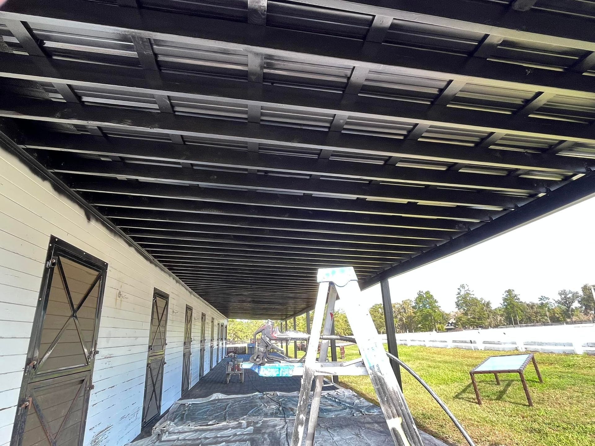 Black metal roof over a white building with several doors. A ladder is in front. Outdoors, sunny day.