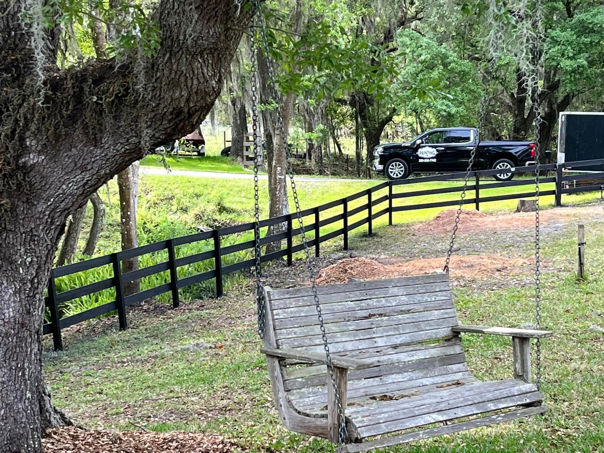 Wooden swing in a yard with a black fence, truck, and trees.