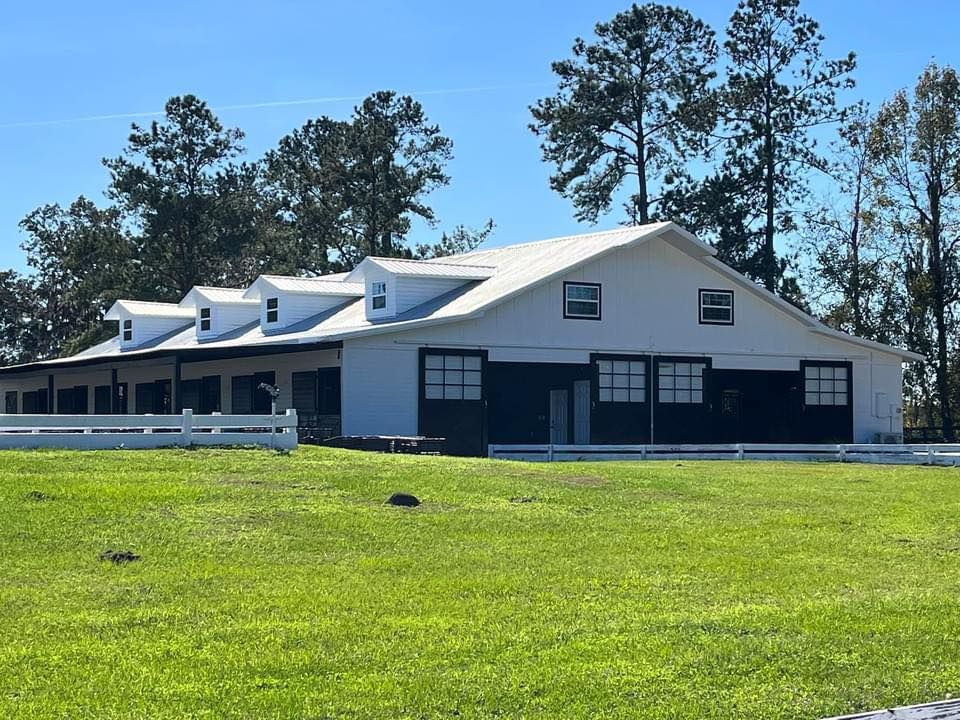 White barn with black accents and a white fence on a grassy field, under a bright sky.