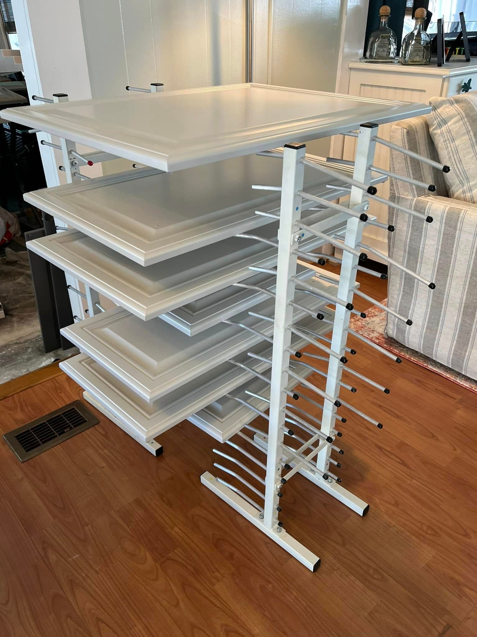 White paint drying rack holding several freshly painted cabinet doors on a hardwood floor.