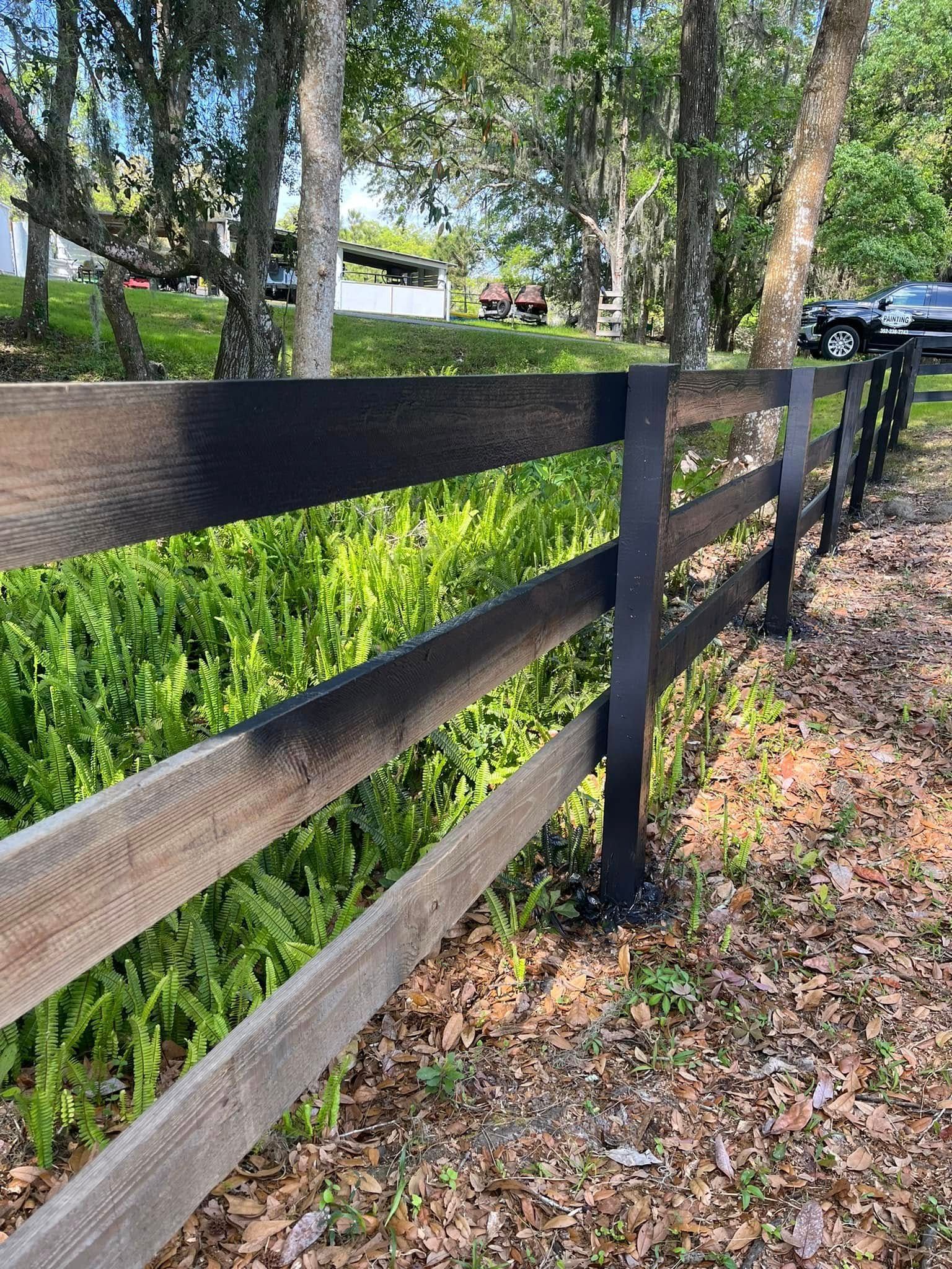 Wooden fence with black posts and three horizontal rails, bordering green foliage.