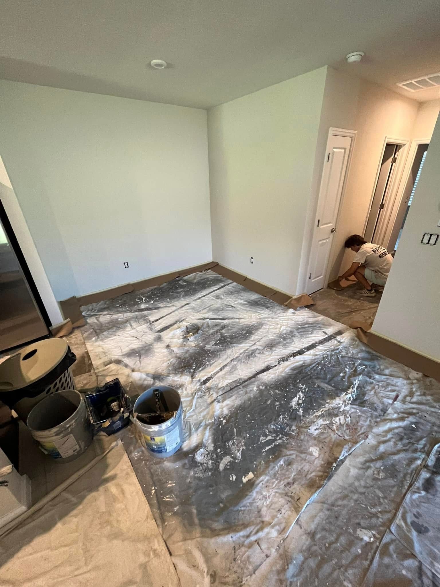 Room being painted; drop cloths on floor, paint buckets, painter working by doorway.