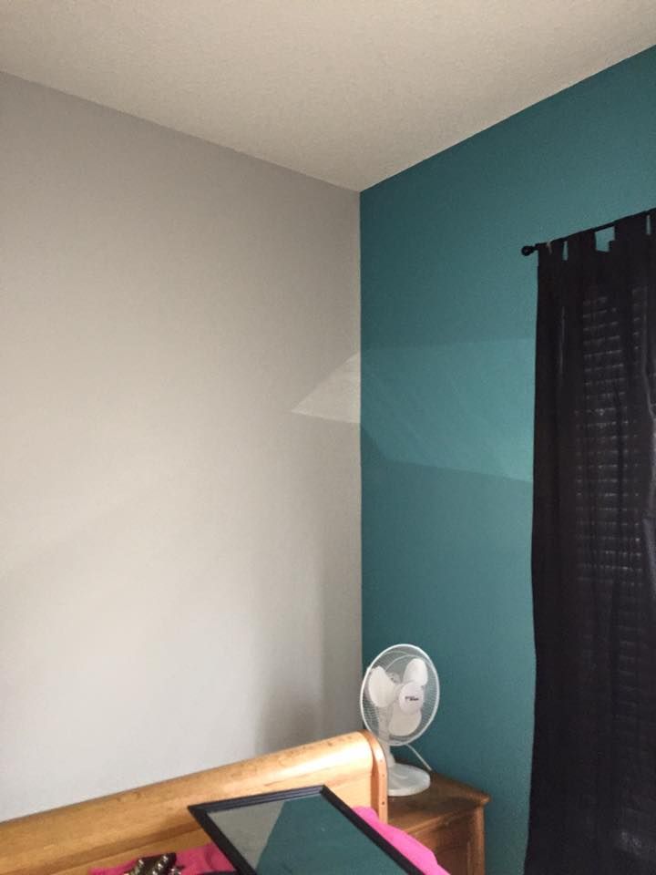 Corner of room with teal and gray walls, white ceiling, black curtains, and a fan on a wooden nightstand.