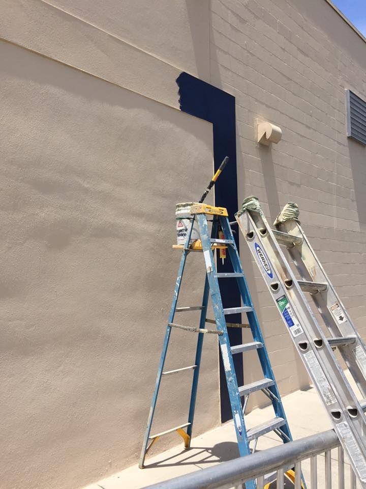 Painting a dark blue vertical stripe on a beige building exterior with a ladder, paint can, and roller.