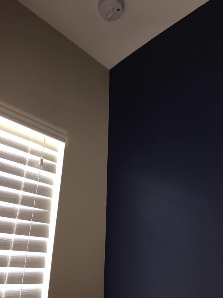 Corner of a room with a window, tan wall, and navy wall. A smoke detector is on the white ceiling.