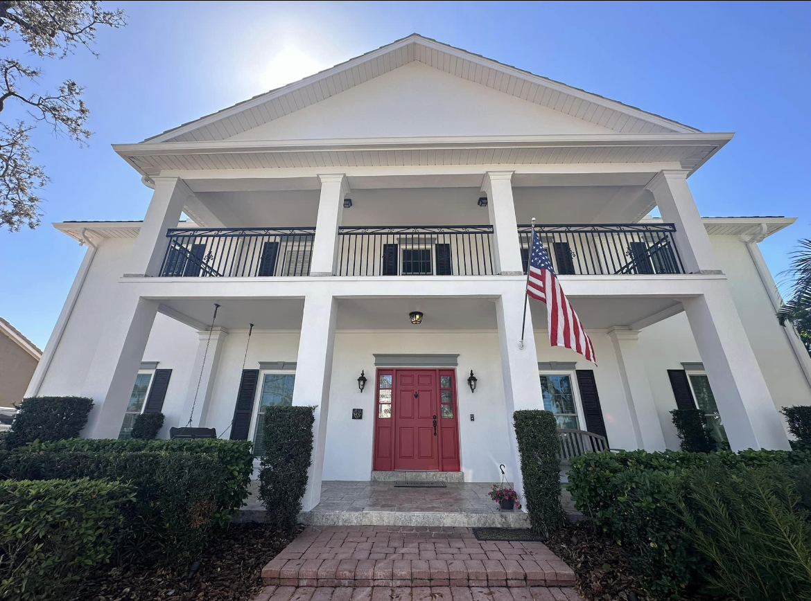 Exterior view of a horse stable with stalls and a long walkway. Black roof and doors, white brick walls, green lawn.