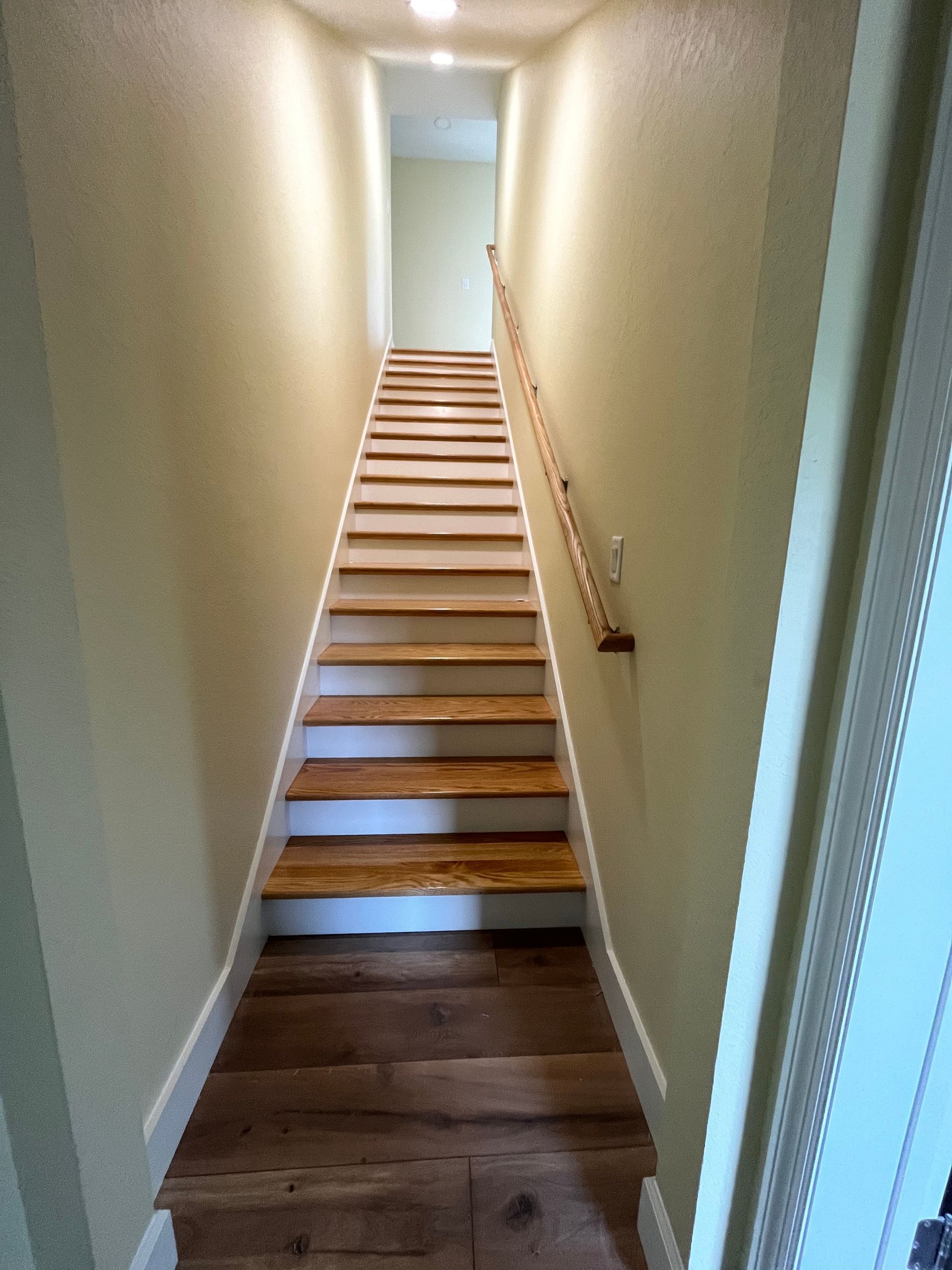 Wooden staircase between light-colored walls, leading upwards. Handrail on the right.