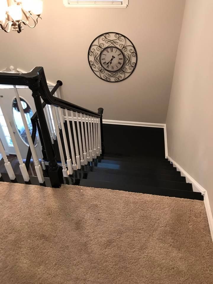 Staircase with black carpet, white spindles, and black railing. A clock hangs on a beige wall.