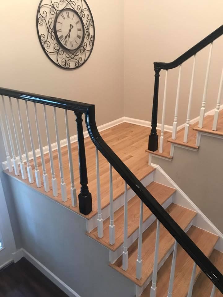 Staircase with light wood steps, white spindles, and black railing. A clock hangs on the beige wall.
