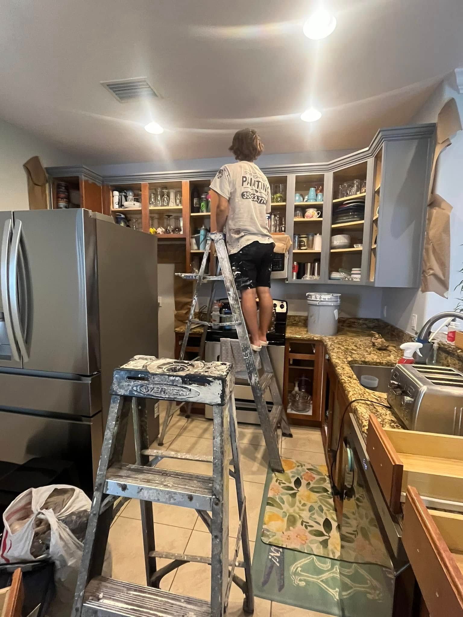 Person on ladder painting kitchen cabinets, room under renovation, with stainless steel appliances.