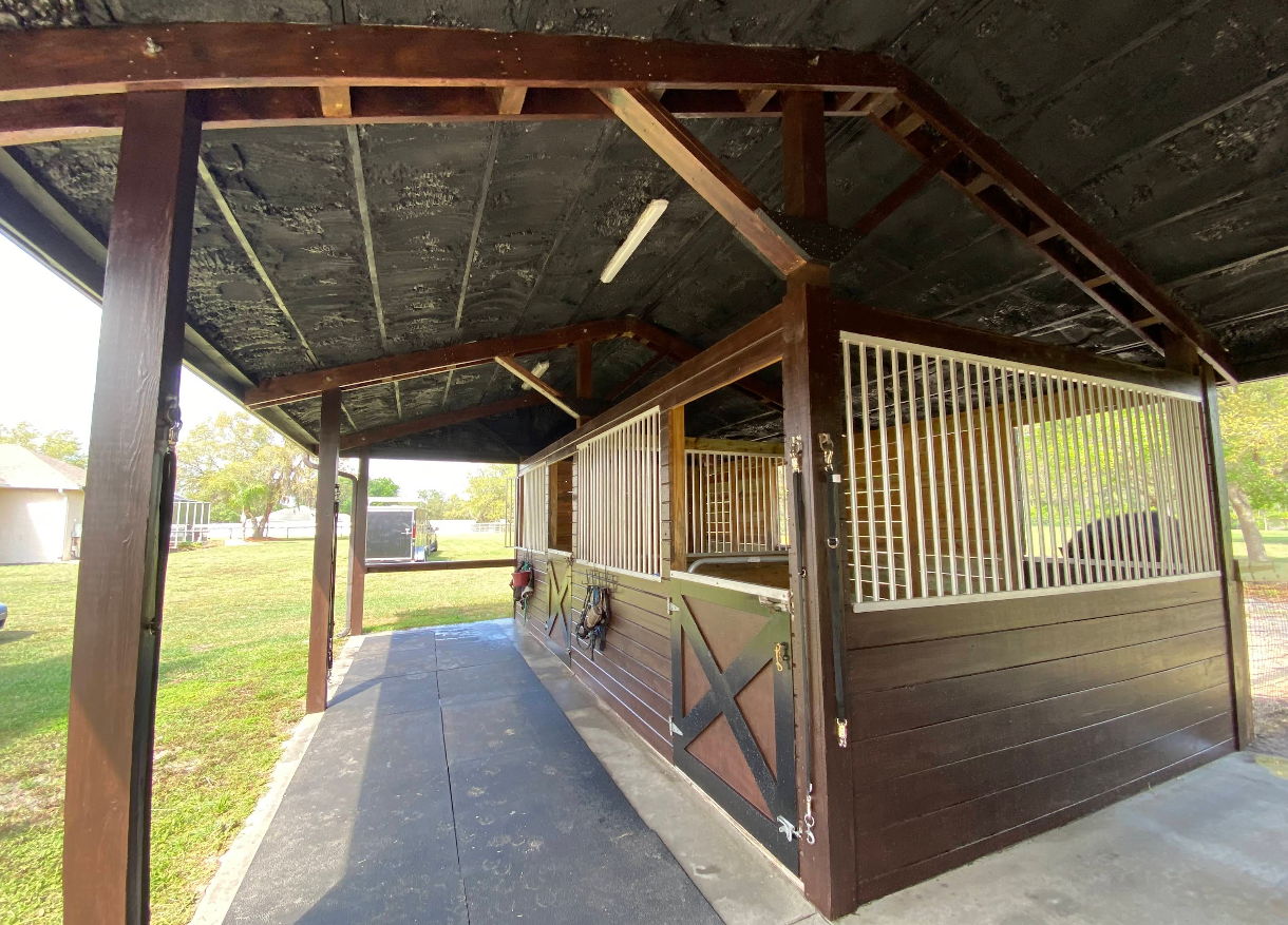 Brown horse stable with an open-air overhang and a paved walkway, set against a grassy yard.