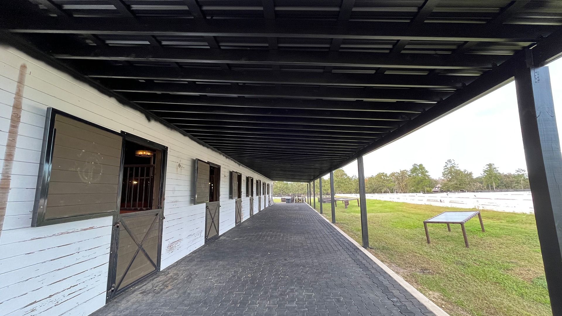 Exterior of a horse stable with stalls and a long walkway. Black roof and doors, white brick. Green grass and trees visible.