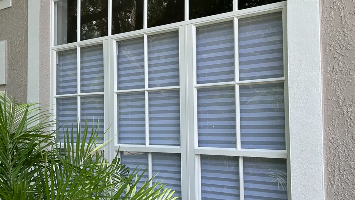 White-framed window with gray pleated blinds, on a stucco building with some green plants in front.