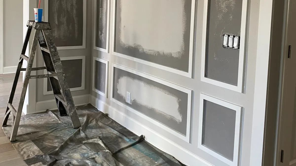 Ladder next to a wall with trim work, partially painted gray and white, with electrical outlets.