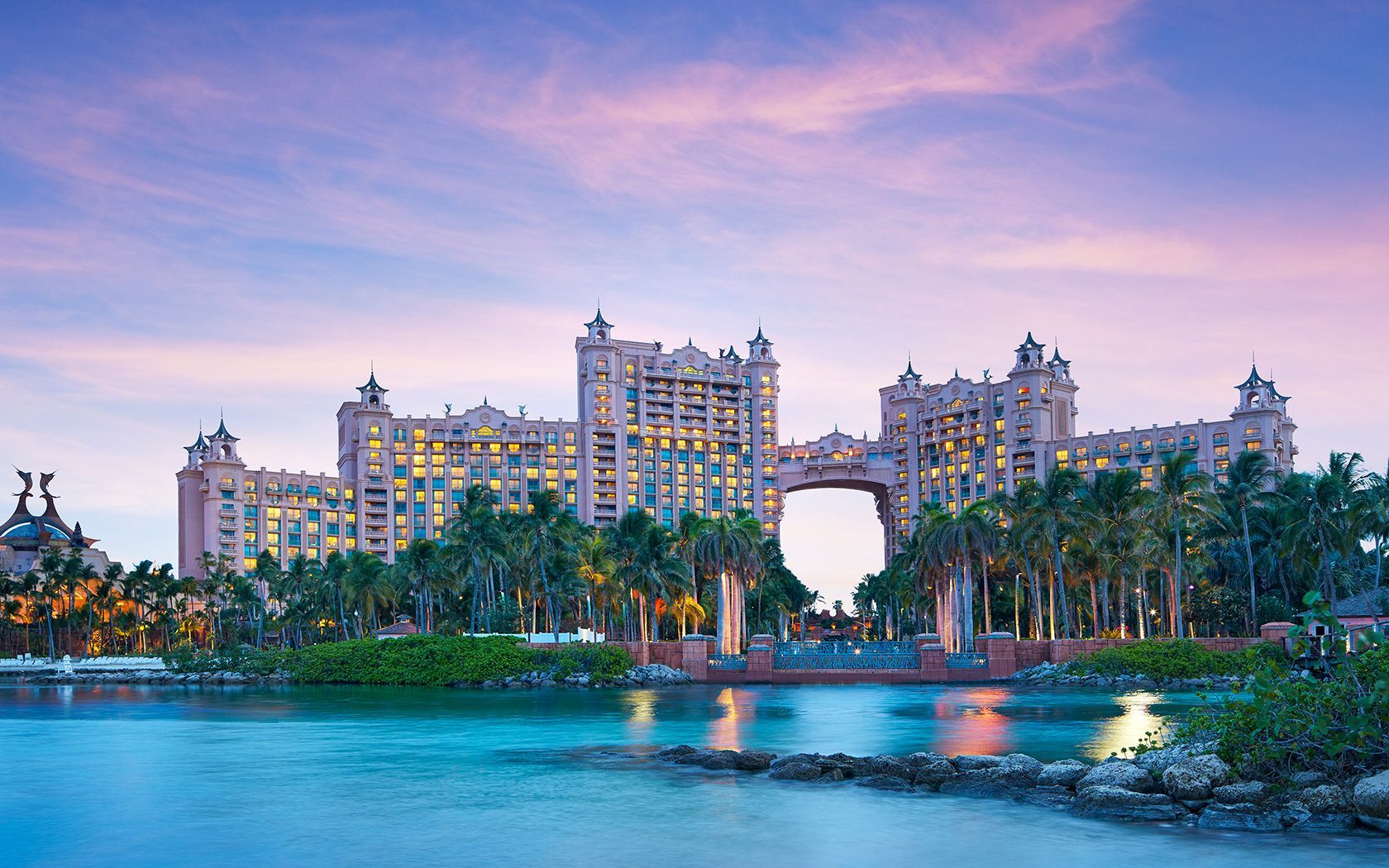 Pink-toned hotel on a tropical waterfront at sunset. Palm trees in front of the building and water in the foreground.