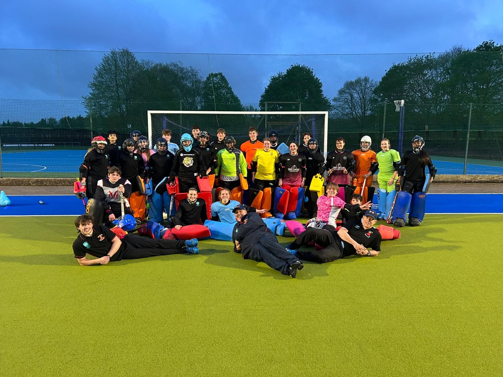 Hockey goalkeepers posing on a field at dusk, with a goal and trees in the background