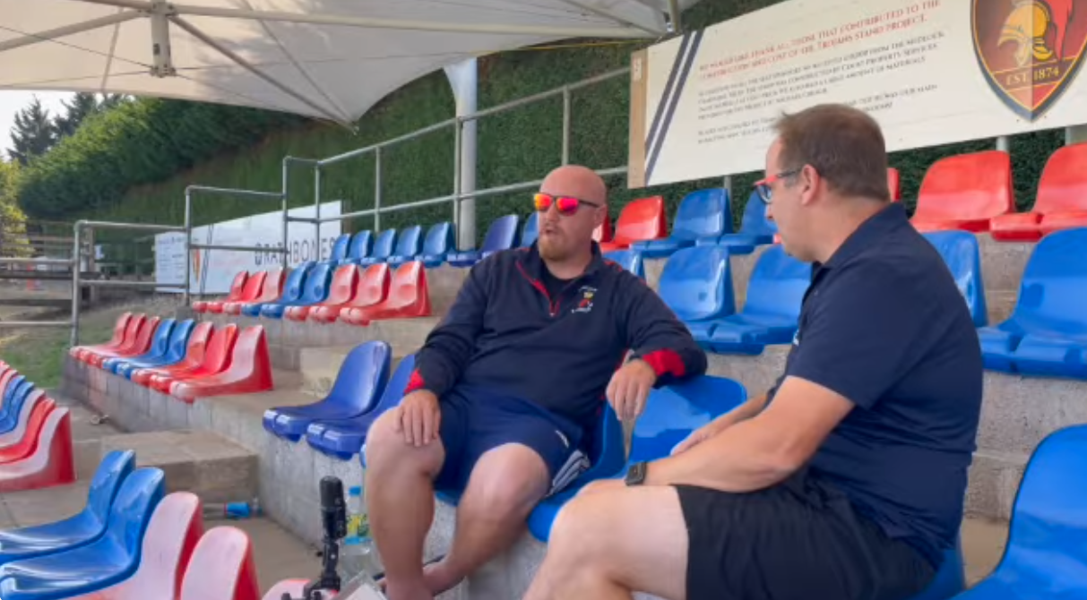 Two men sit on blue and red stadium seats, discussing something outdoors. One wears red sunglasses, the other glasses.