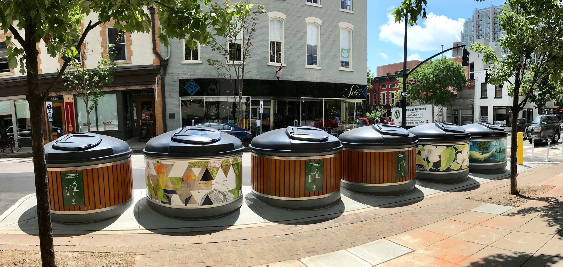 Six decorative trash receptacles lined up on a sidewalk. Buildings in the background. Bright sunny day.