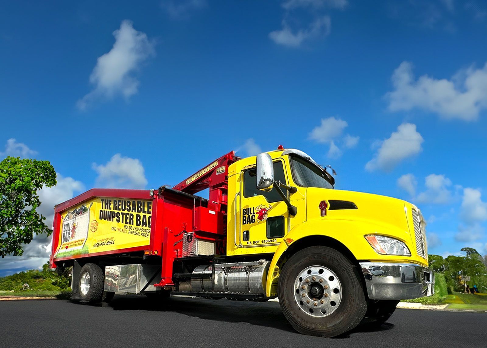 Yellow and red dumpster truck parked on asphalt under a blue sky with clouds.