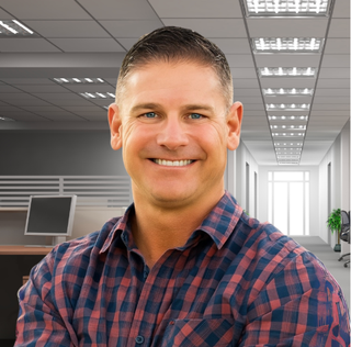 Man smiling, wearing a plaid shirt, in an office setting with desks and a hallway.