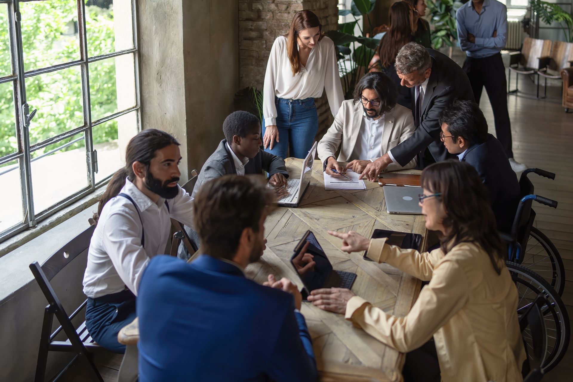Business meeting around a large table; people working on laptops, looking at documents, in an office with large windows.