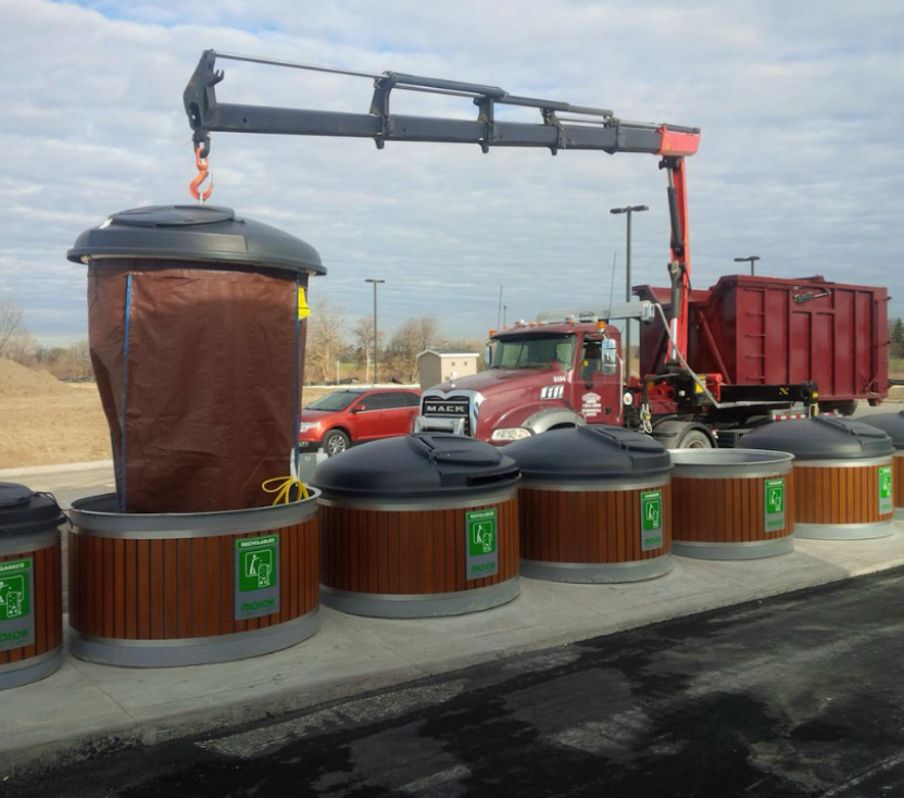 A crane lifting a brown waste bag out of a public trash container, with a red truck in the background.