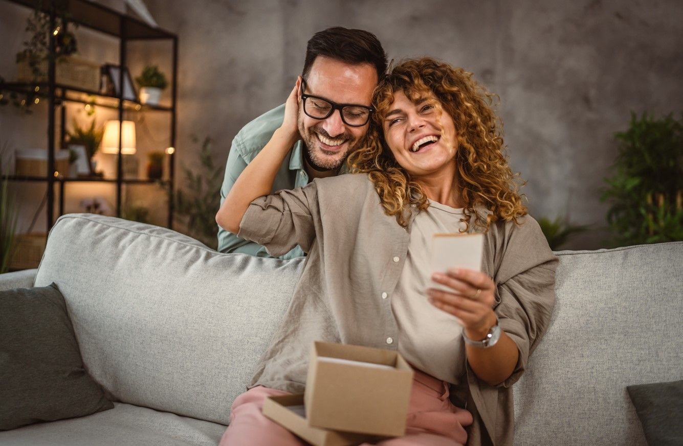 Woman opening gift box, smiling. Man hugs her from behind, both laughing. Interior, living room.