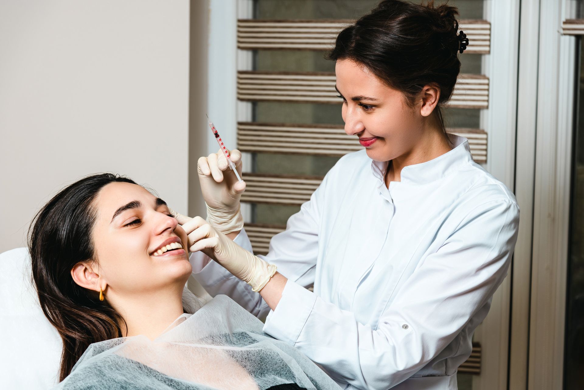 Person receiving a cosmetic injection near the nose by a smiling medical professional. Indoors.