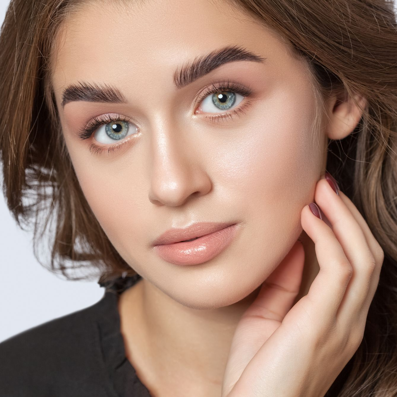 Woman with light skin, blue eyes, and brown wavy hair, wearing pink eyeshadow, gently touching her cheek.