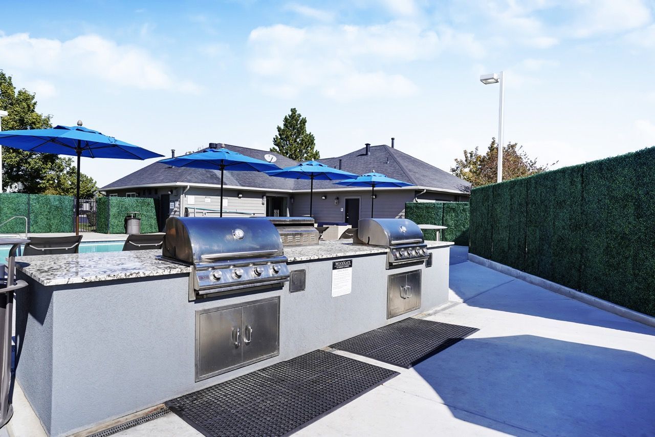 Outdoor community barbecue area at Woodgate with stainless grills, granite counters, and blue umbrellas.