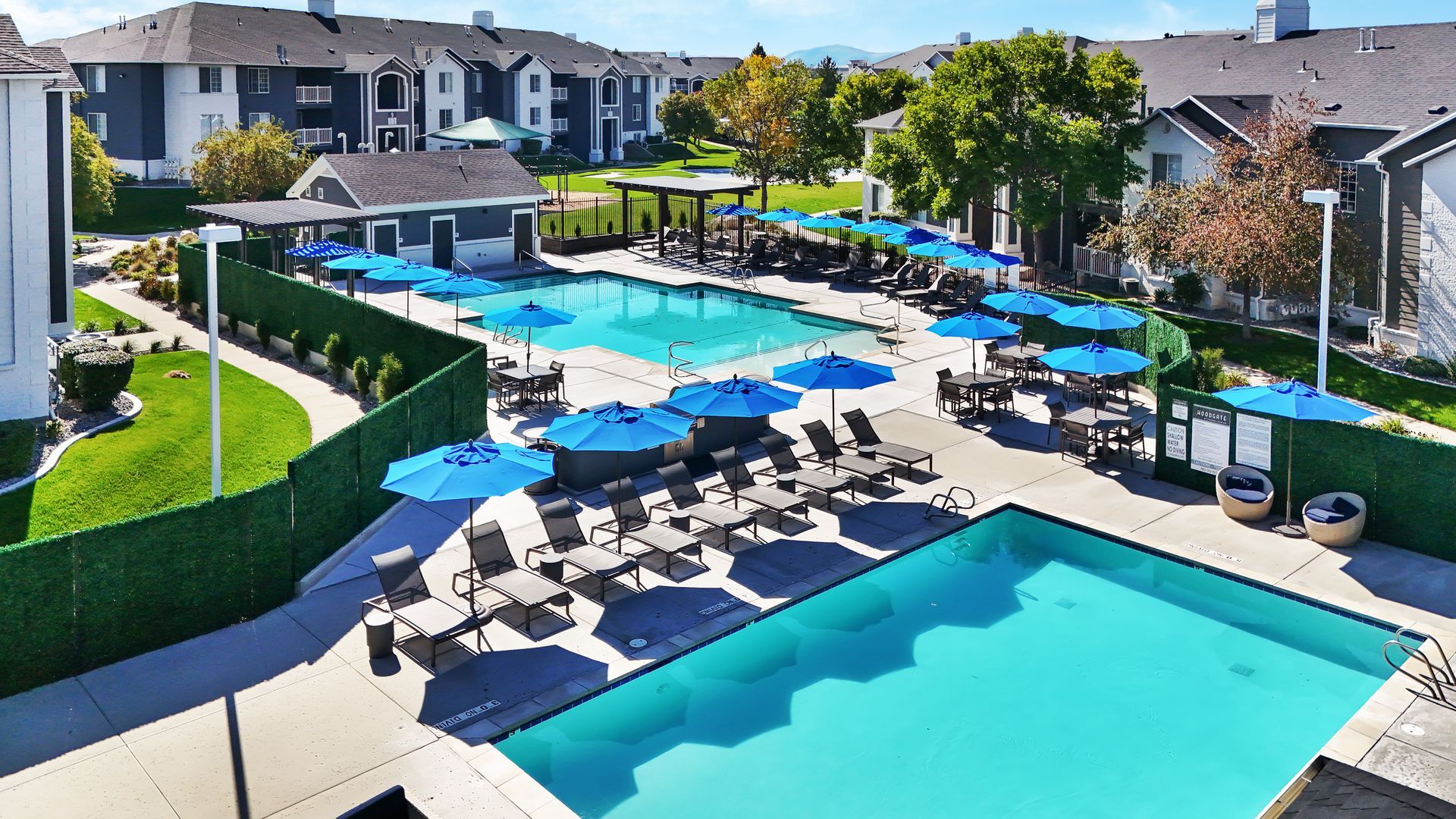 Aerial view of a community pool area at Woodgate with blue umbrellas and lounge chairs among apartment buildings.