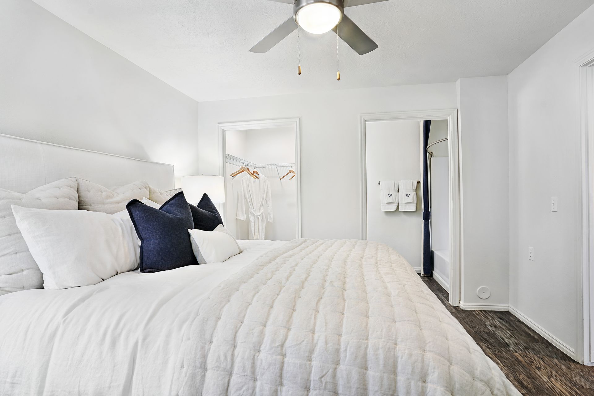 Bedroom in a modern apartment with white bedding, navy pillows, a ceiling fan, and closet doors at Woodgate.