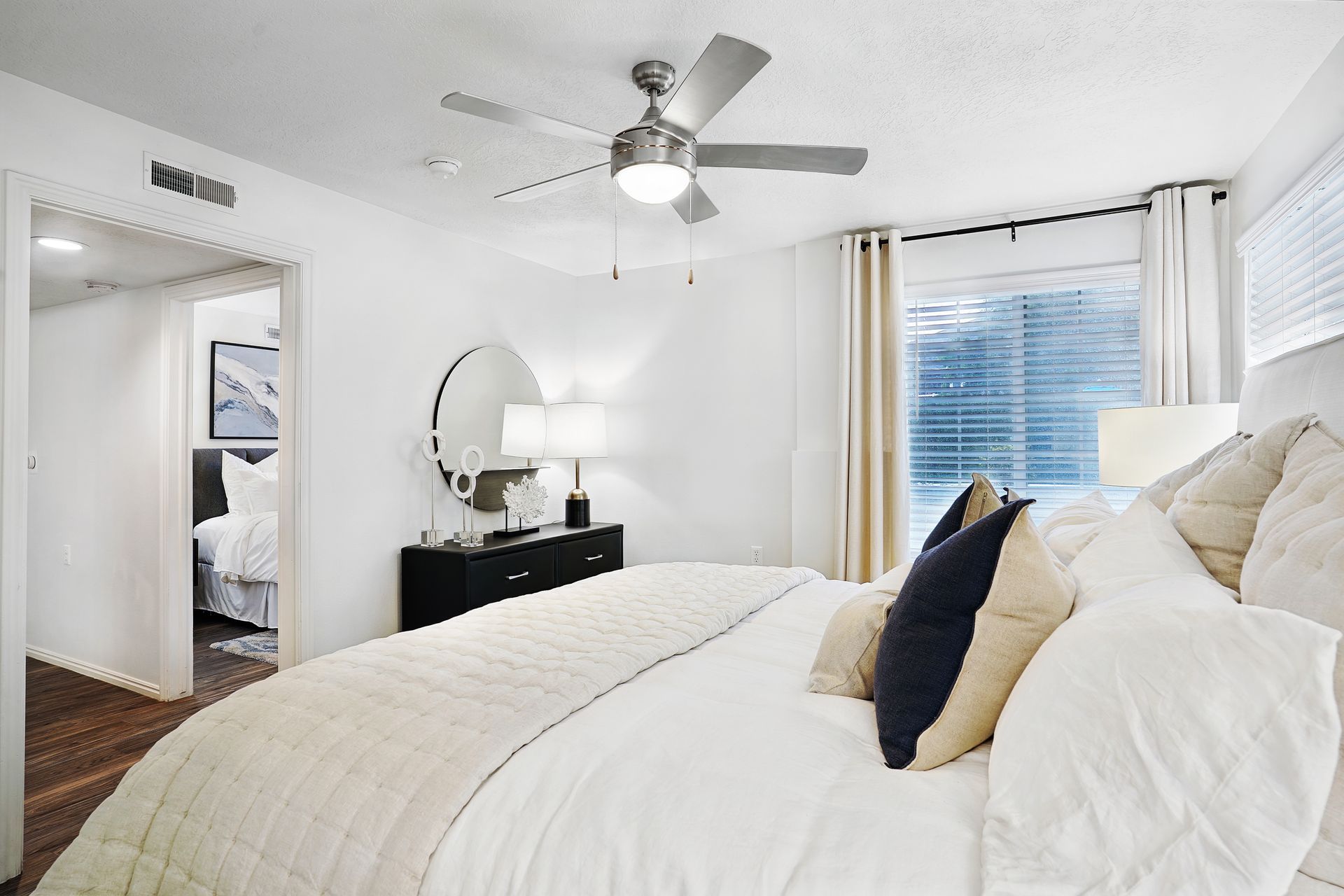 Bright, modern bedroom in a white-walled apartment with a large bed, round mirror, and window at Woodgate.