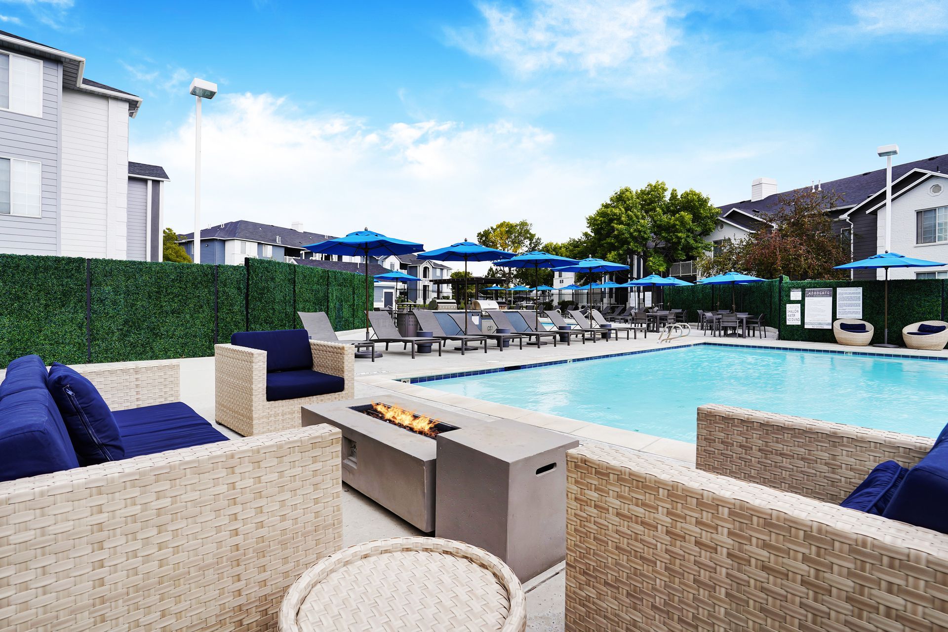 Outdoor apartment pool area at Woodgate with blue umbrellas, lounge chairs, and woven seating.