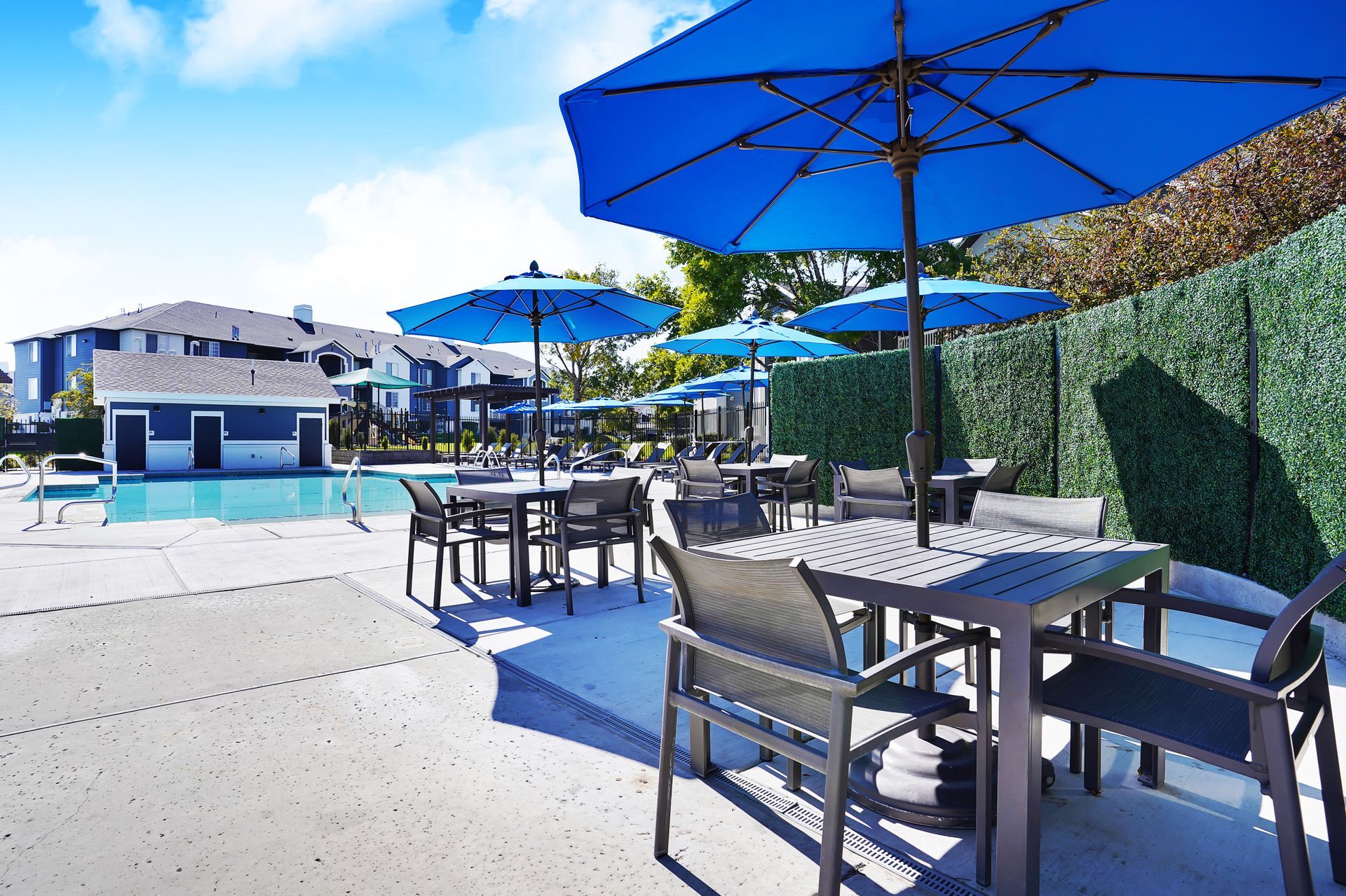 Outdoor community pool area at Woodgate with blue umbrellas, tables and chairs on a concrete deck.