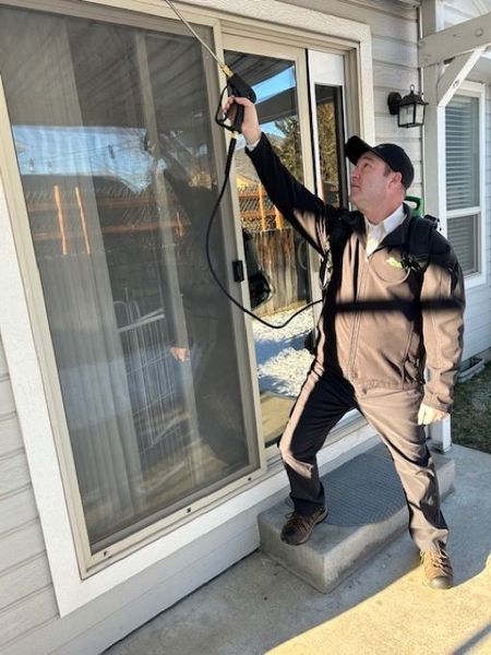 A pest control technician in a uniform uses a spray wand to treat the exterior frame of a sliding glass door.
