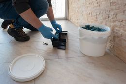 A person wearing blue gloves places blue bait blocks into a black pest control station next to a white bucket on a floor.