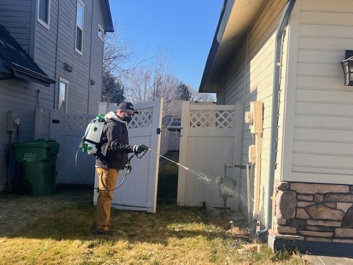 A pest control technician in a black jacket and tan pants sprays liquid near the base of a house's exterior.
