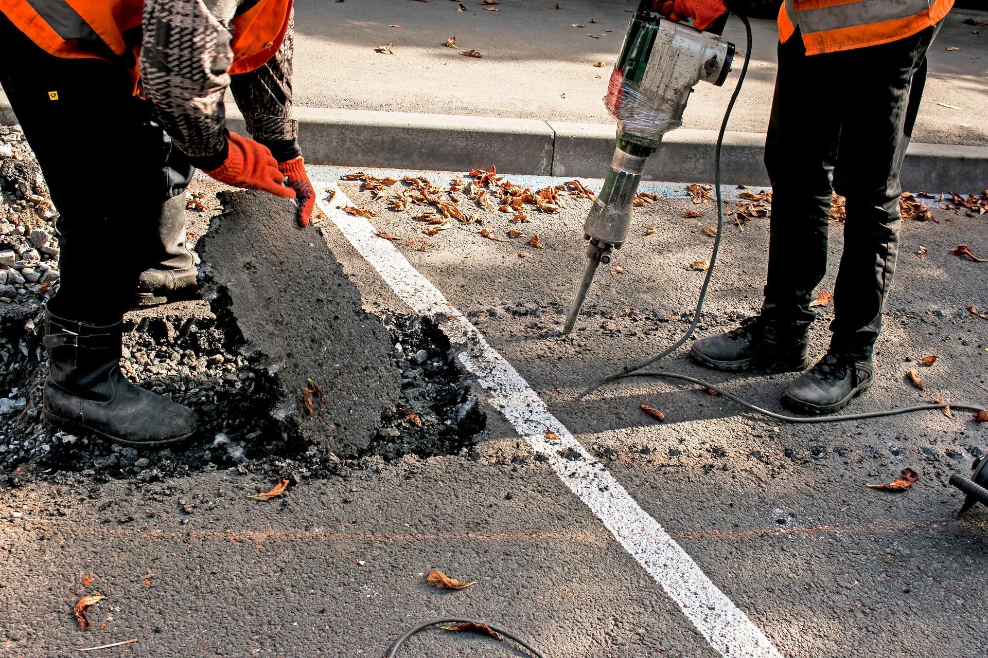 Two workers removing a white road marking with a jackhammer on an asphalt road.